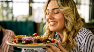 Uśmiechnięta blondynka delektuje się aromatem grillowanego mięsa z warzywami podanego na jasnym talerzu w restauracji
