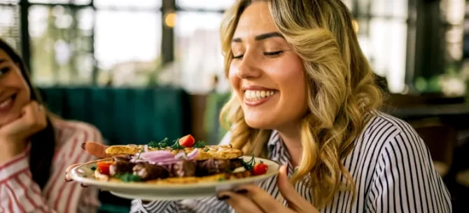 Uśmiechnięta blondynka delektuje się aromatem grillowanego mięsa z warzywami podanego na jasnym talerzu w restauracji