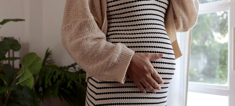 Czy odra jest niebezpieczna w ciąży? Możliwe powikłania Przyszła mama w beżowym swetrze i sukience w paski czule dotyka swojego zaokrąglonego brzucha przy jasnym oknie, zbliżenie na brzuch