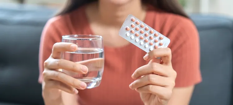 Tabletki antykoncepcyjne w czasie karmienia piersią – jakie można stosować? Dłoń trzymająca szklankę z przejrzystym płynem oraz blister leków hormonalnych na tle pomarańczowej koszulki