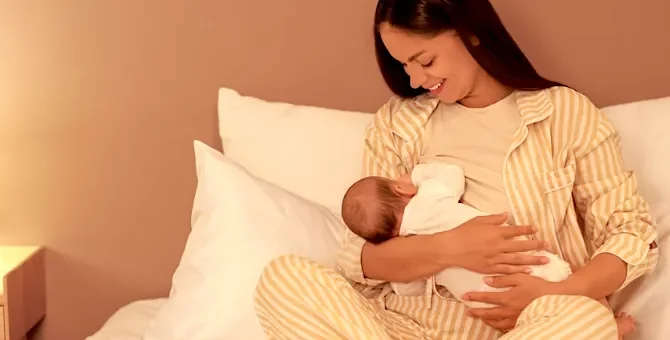 Uśmiechnięta kobieta w pasiastej piżamie karmi w łóżku wieczorem piersią noworodka, na tle oświetlonej lampką nocną ściany