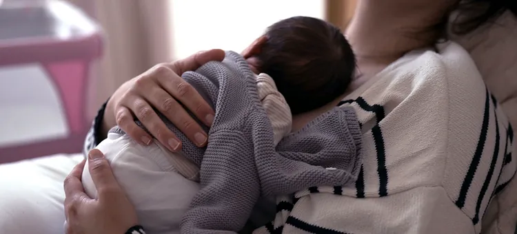 Karmienie piersią a więź mamy i dziecka – czy ma znaczenie? Kobieta w jasnym swetrze obejmuje śpiące niemowlę ubrane w szary kardigan