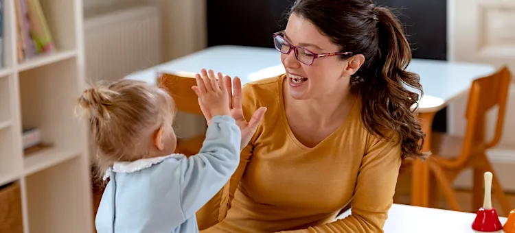 Radosna opiekunka w musztardowej bluzce bawi się z maluchem przy jasnym stoliku