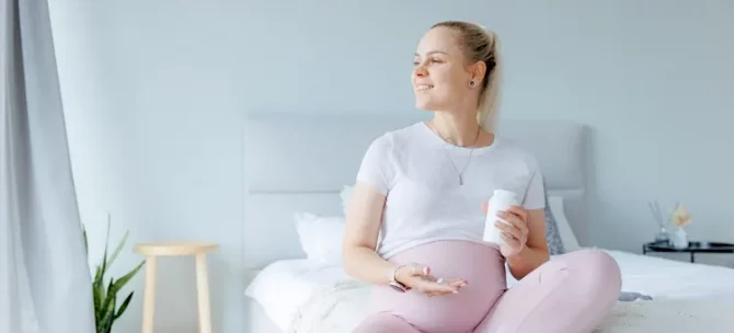 Uśmiechnięta ciężarna trzymająca suplementy diety w jasnej sypialni