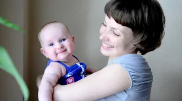 Radosne dziecko w niebieskim ubranku w objęciach uśmiechniętej kobiety na tle szarej ściany