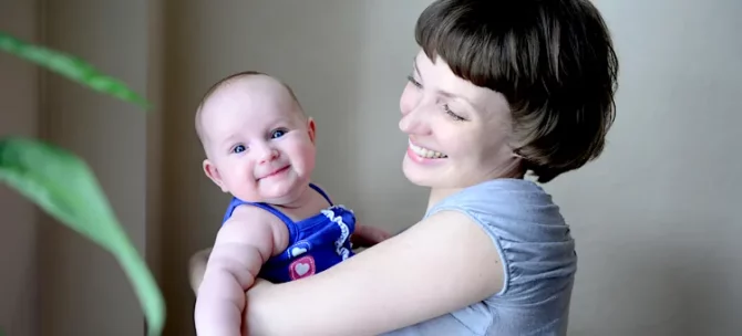 Radosne dziecko w niebieskim ubranku w objęciach uśmiechniętej kobiety na tle szarej ściany
