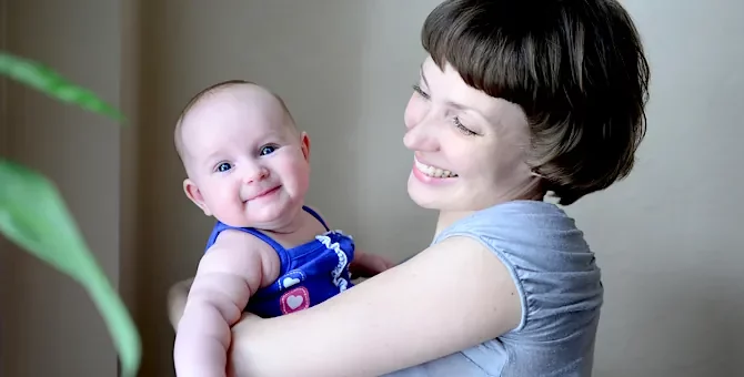 Radosne dziecko w niebieskim ubranku w objęciach uśmiechniętej kobiety na tle szarej ściany